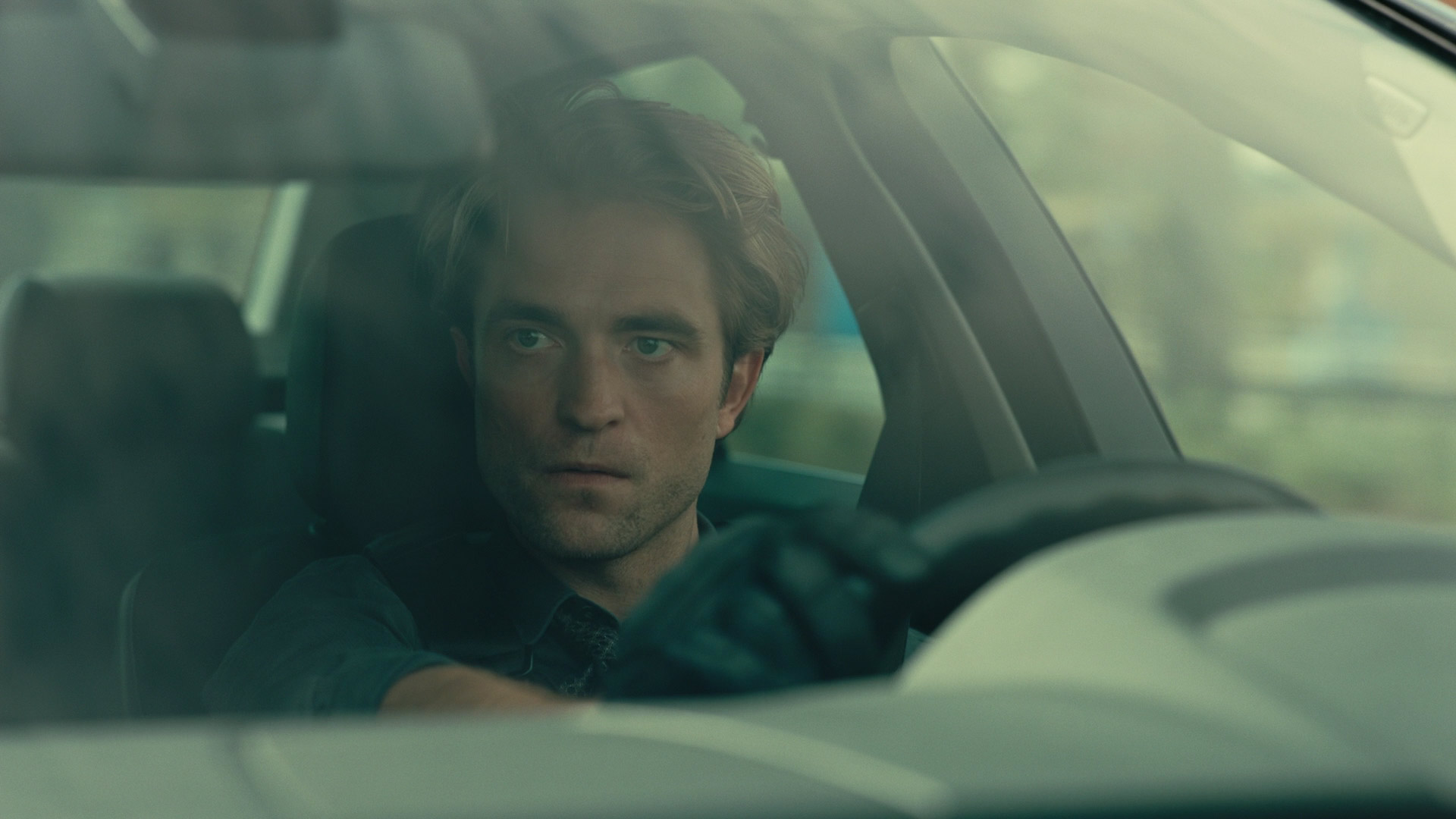 a man with short, blonde hair is driving a car. he is wearing a dark blue shirt and black leather driving gloves. the image is taken from the perspective of someone outside the car, looking in through the windshield. the lighting is subdued, suggesting an overcast day. the overall tone of the image is serious and contemplative.