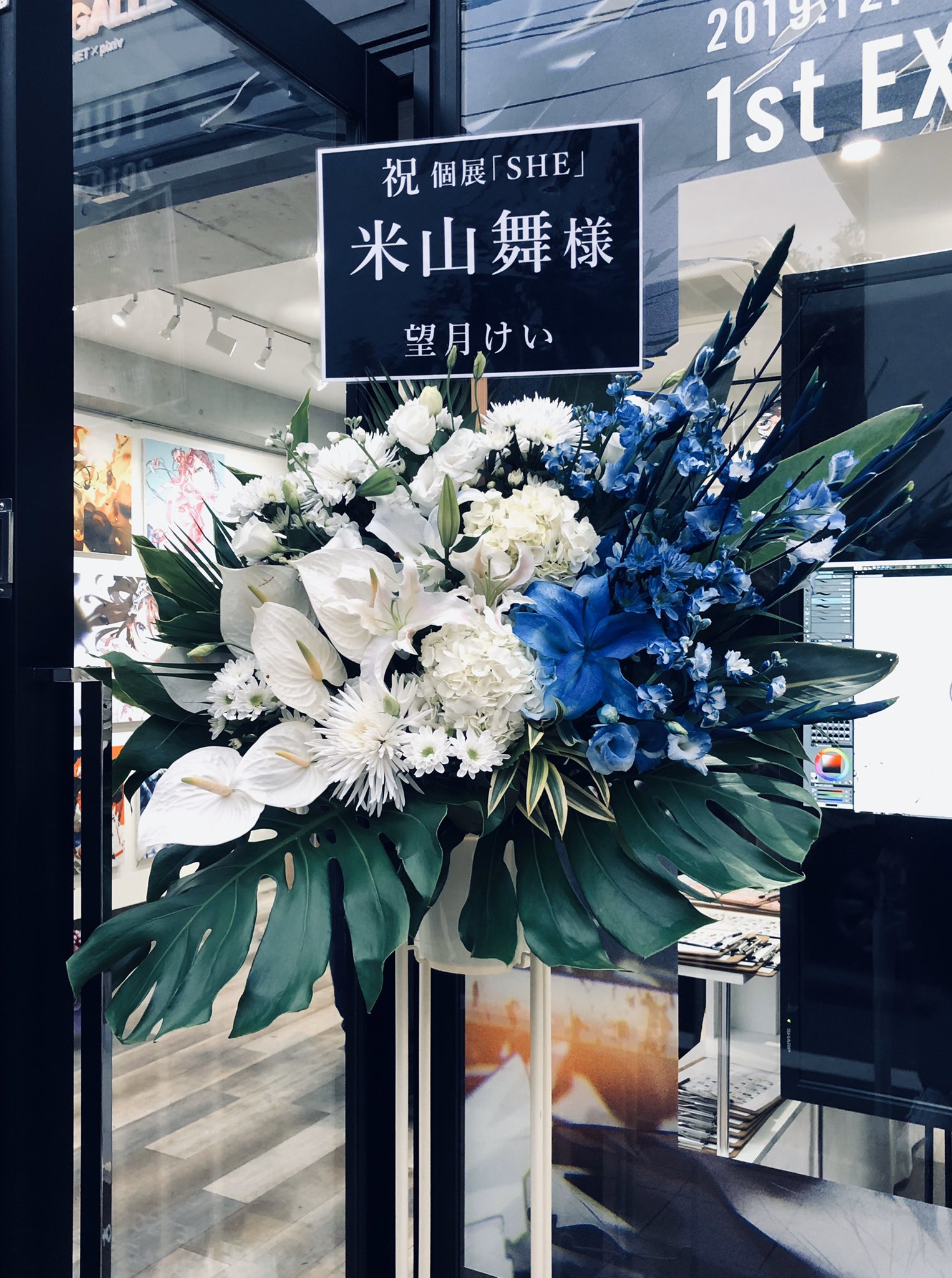 a vibrant floral arrangement stands in front of a glass storefront. the arrangement features white lilies, blue delphiniums, and lush greenery. a black sign with white japanese characters congratulates an artist named "米山舞" on their solo exhibition titled "she."  the storefront reflects the street and buildings across from it.