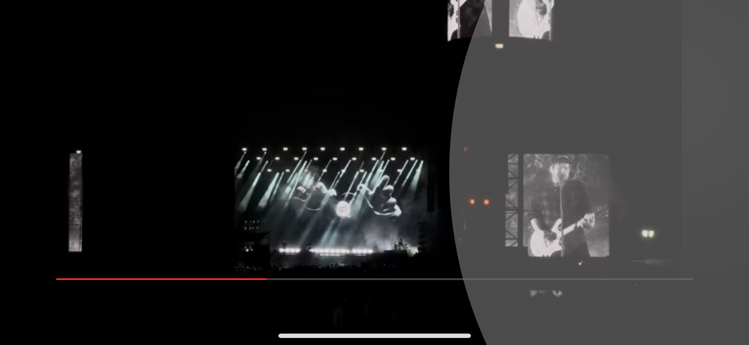 a concert venue with a large stage lit by many spotlights. the stage is silhouetted by a large screen showing the band performing, with the outline of the lead guitarist clearly visible. the crowd is not visible but can be seen in front of the stage. the atmosphere is loud, energetic, and captivating.