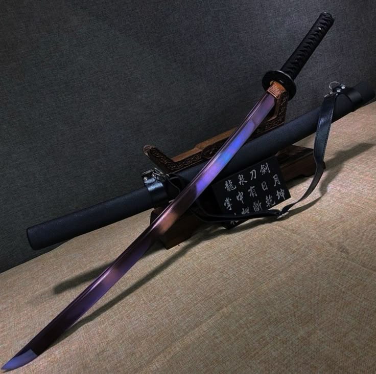 a katana sword with a purplehued blade is laid diagonally across its black scabbard, which rests on a wooden stand. a leather strap is attached to the scabbard. a small plaque with japanese writing sits near the base of the stand. the background is a textured wall.
