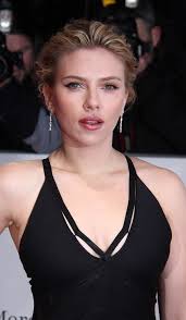 a closeup portrait of scarlett johansson, a renowned hollywood actress. she is elegantly dressed in a black, lowcut dress with thin straps, and is wearing delicate earrings. her blonde hair is styled neatly, and her makeup enhances her features. the setting appears to be a formal event, possibly a red carpet or awards ceremony, as suggested by the blurred background showing other people and what seems to be photographic equipment.  the overall impression is one of glamour and sophistication.