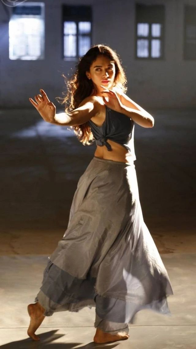 a young woman in a long flowing grey skirt is dancing in a studio. she is posed with her back to the camera and her right arm extended. she is in midpose, her skirt flowing around her.  the studio is lit with spotlights and the background is blurred.