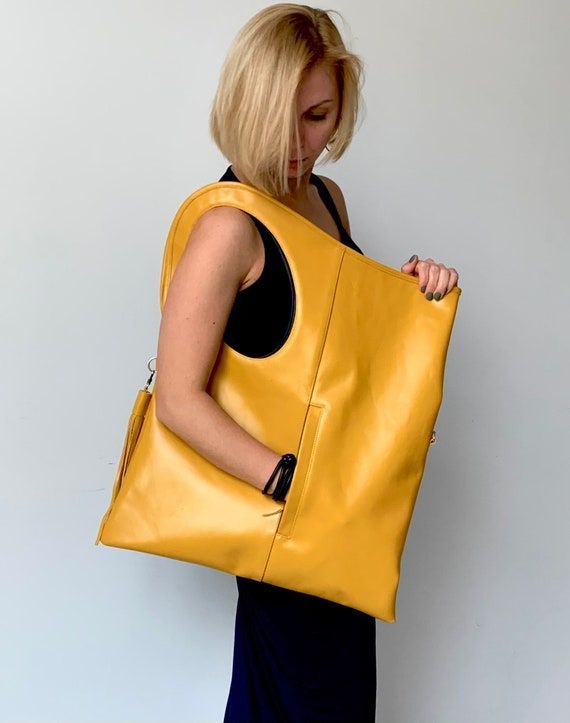 a woman with blonde hair is holding a large yellow leather bag. she is wearing a sleeveless black dress. the bag is a large tote style with a cutout handle and a small pocket on the front. the background is a plain white wall.