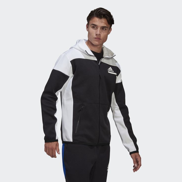 a man is wearing a black and white adidas zipup hoodie. he's wearing the hoodie with the hood down. the adidas logo is visible on the right chest of the jacket. he is wearing black pants. the man is looking straight into the camera. he has short brown hair and a light beard. the background is white.
