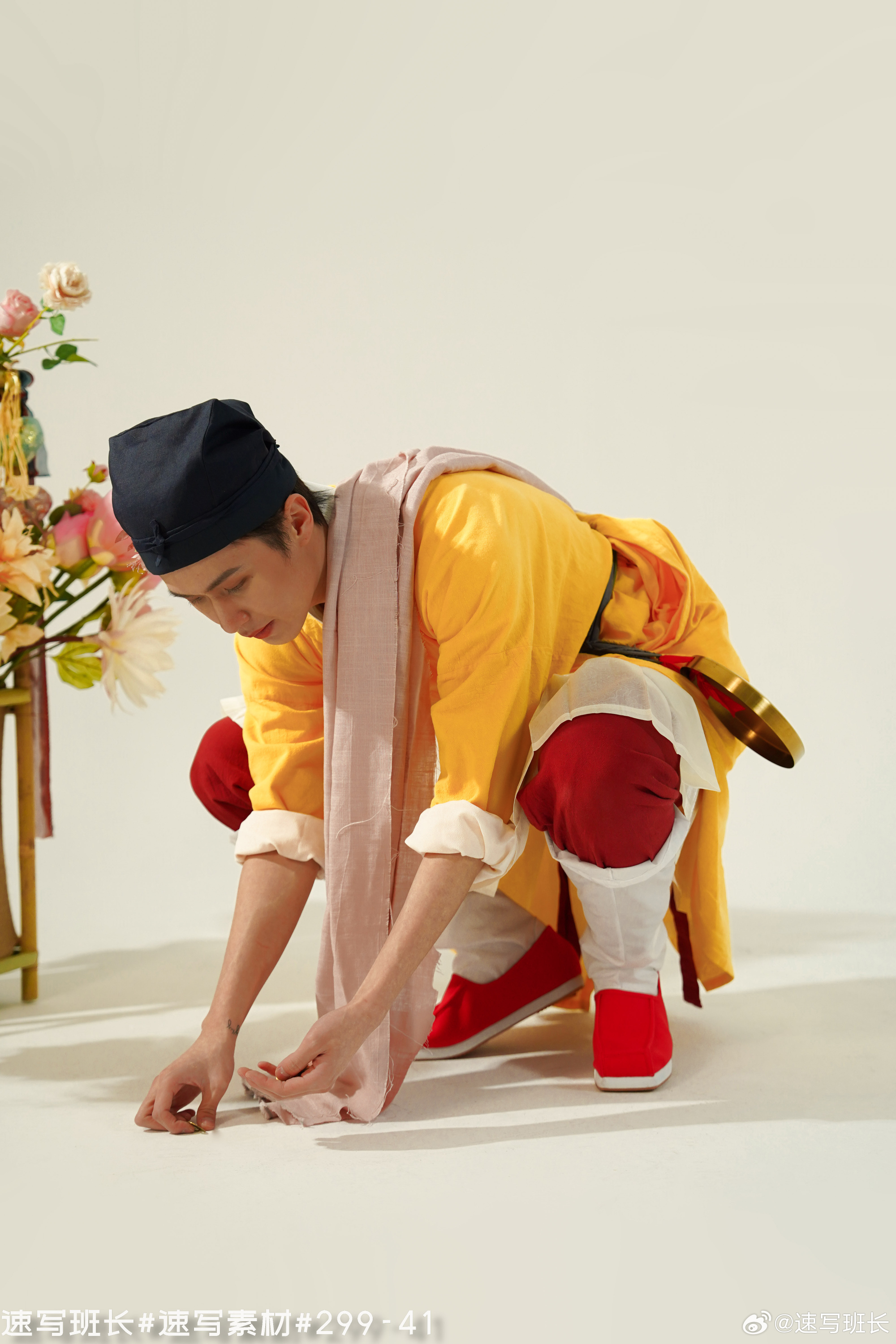 a man in bright, traditional chinese clothing kneels on the ground, his hand reaching down as if picking up a small object. the lighting is soft and diffused, creating a warm and inviting atmosphere. to his left sits a stand with a vibrant floral arrangement. the image evokes a sense of cultural heritage and performance art.