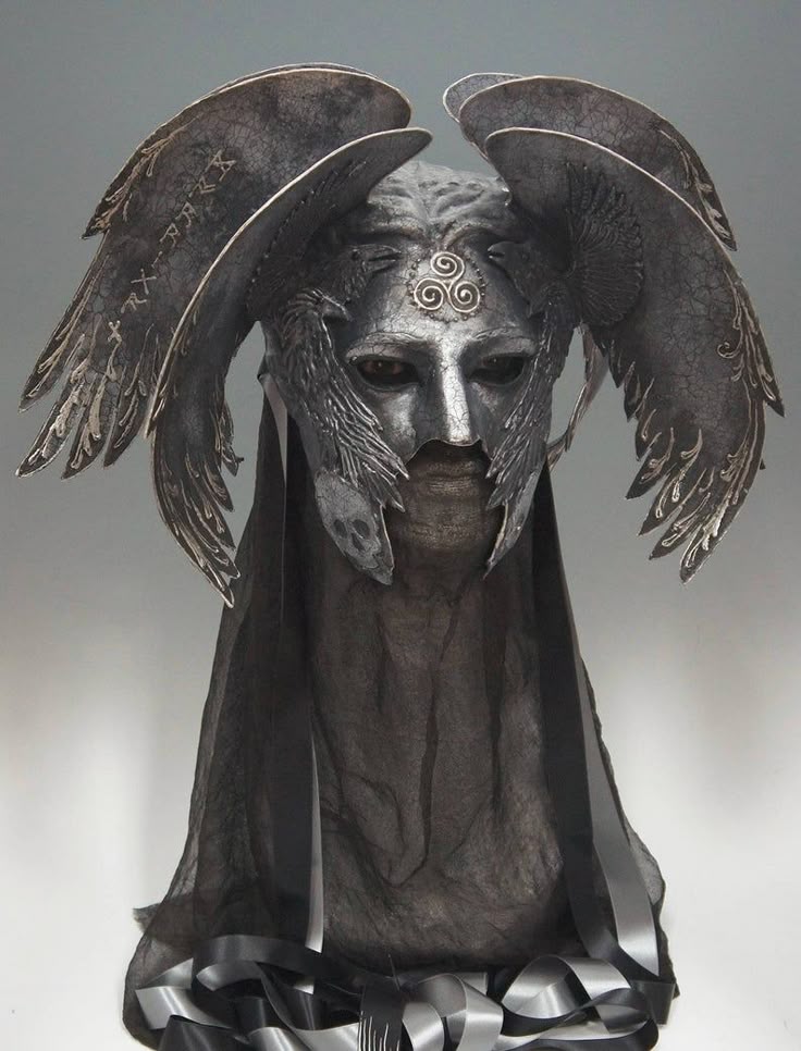 a silver mask sits atop a figure draped in a dark veil. the mask features wings extending upwards and outwards from the sides, with runes inscribed on them. two ravenlike birds adorn the mask near the cheeks. a celtic triple spiral symbol is centered on the forehead of the mask. the figure is draped in a dark, sheer veil, with a collection of black and silver ribbons gathered at the base. the overall appearance suggests a fantasy or gothic aesthetic.