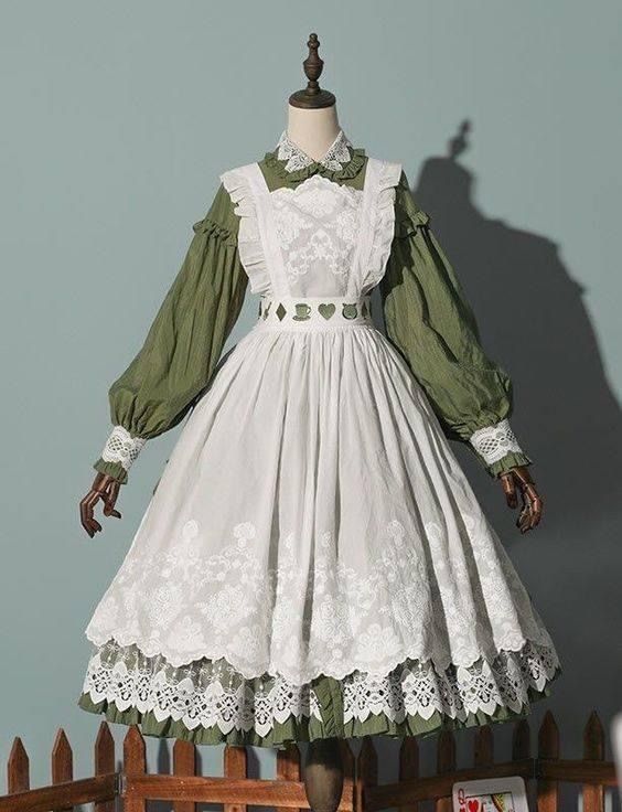 the image shows a white and green lolita fashion dress. it is a longsleeved dress with a high waistline and a full skirt. the dress has an apronlike overlay with floral embroidery and lace detailing. the dress is displayed on a mannequin against a blue wall with a wooden fence in the foreground.