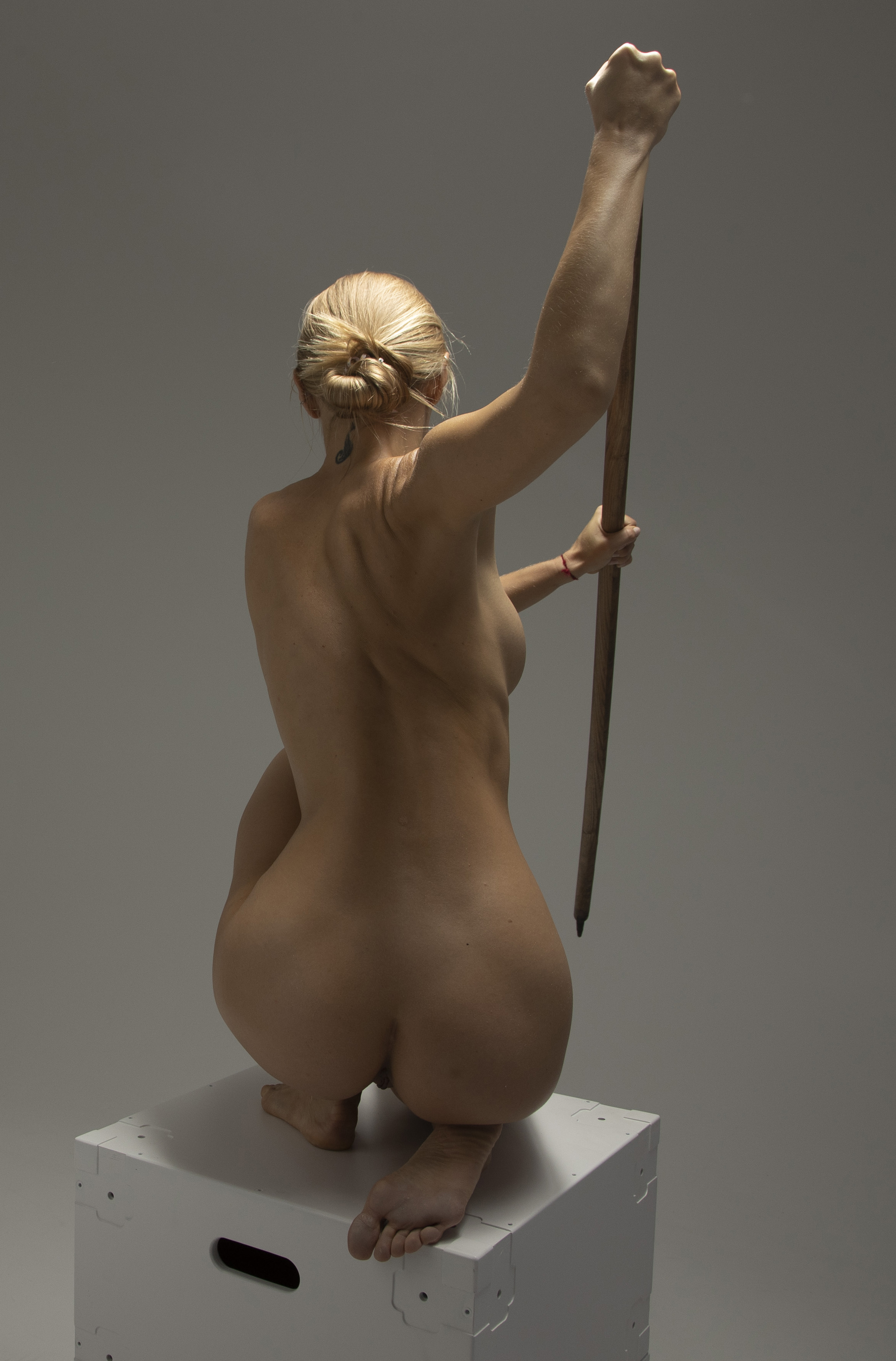 a nude woman with blonde hair in a bun is squatting on a white box. she is facing away from the camera and holding a brown pole in her right hand, which is raised above her head. the background is a neutral gray.