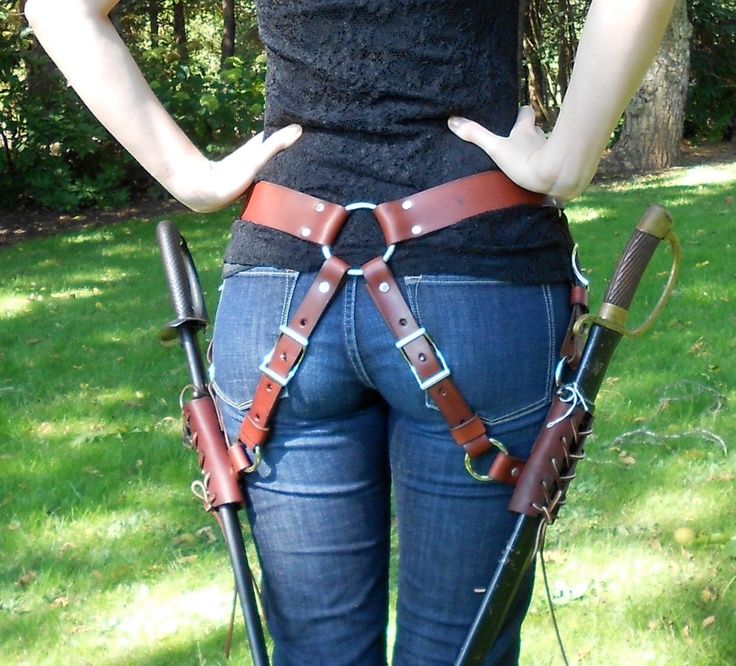 a woman is shown from the back, wearing blue jeans, a black top, and a leather harness around her waist and thighs. two swords, possibly katanas, are hanging from the harness on either side of her hips. she is standing in a grassy outdoor area with trees in the background. her hands are on her hips.