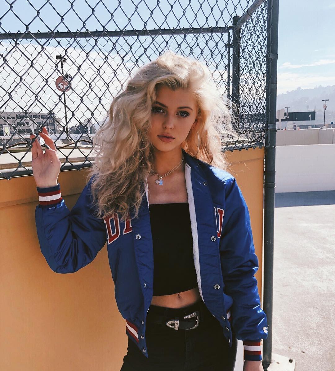 a woman with long blonde curly hair stands outside wearing a black tube top, black jeans, and a blue varsity jacket. she is accessorized with a belt and necklace. she leans against an orange wall next to a chain link fence with a parking garage in the background.