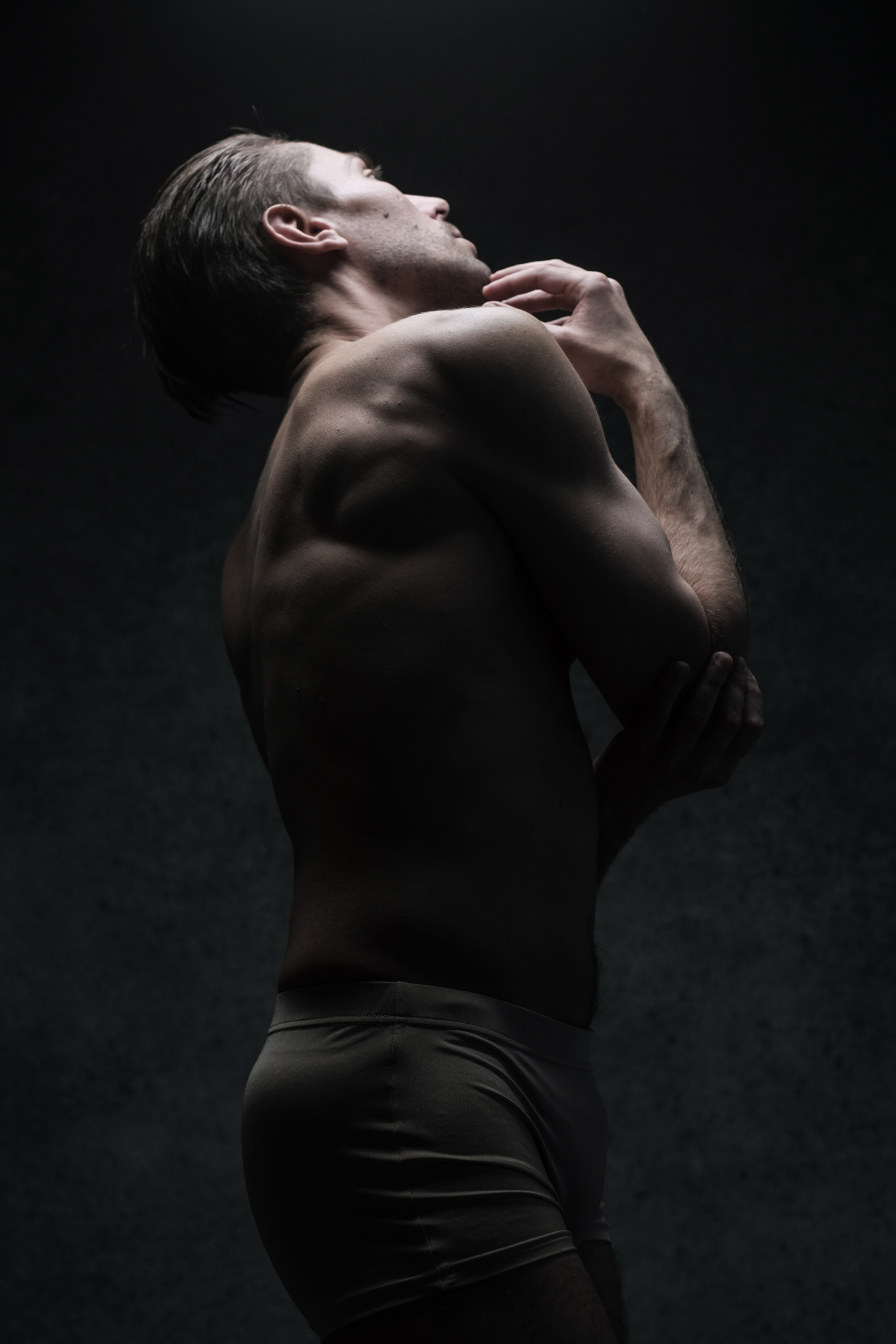 the image showcases a shirtless muscular man in underwear against a dark studio background. he is dramatically lit with a single light source from above, highlighting his physique and creating a chiaroscuro effect. the man's pose is contemplative, with his head tilted back and his eyes closed.