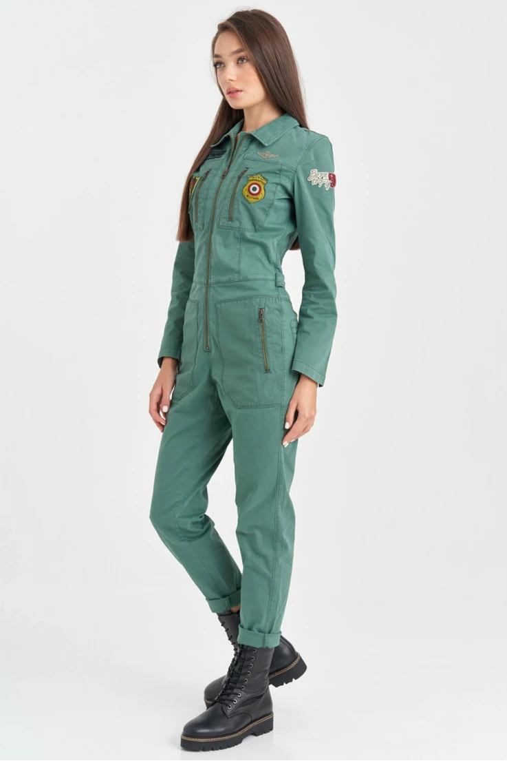 a woman wearing a green jumpsuit and black boots stands in a studio. she has her hair pulled back off her shoulders and her arms are by her side. she is looking at the camera.