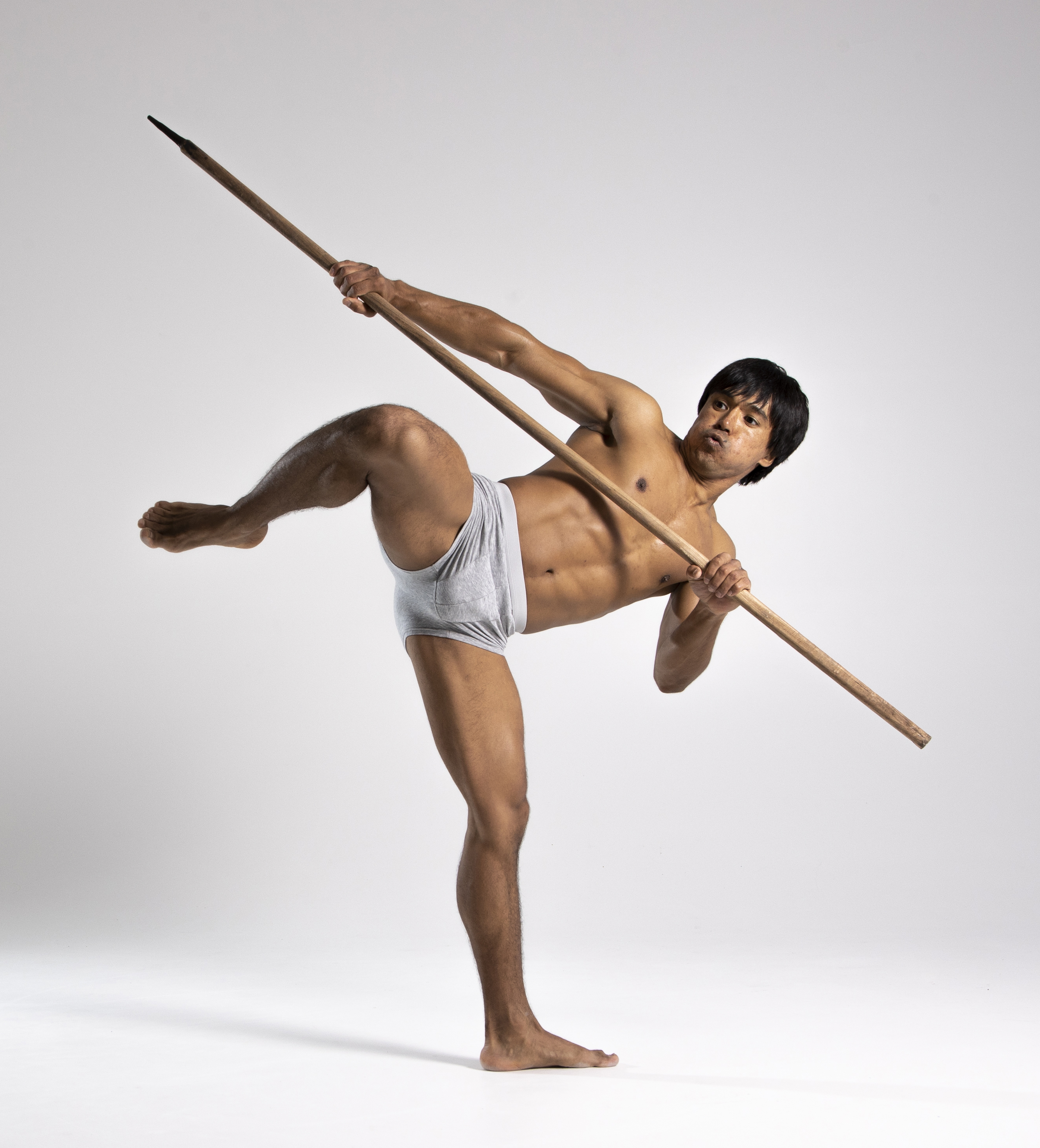 a shirtless man wearing gray underwear is holding a spear while balancing on one leg in a martial arts pose against a white background. he is muscular and athletic.