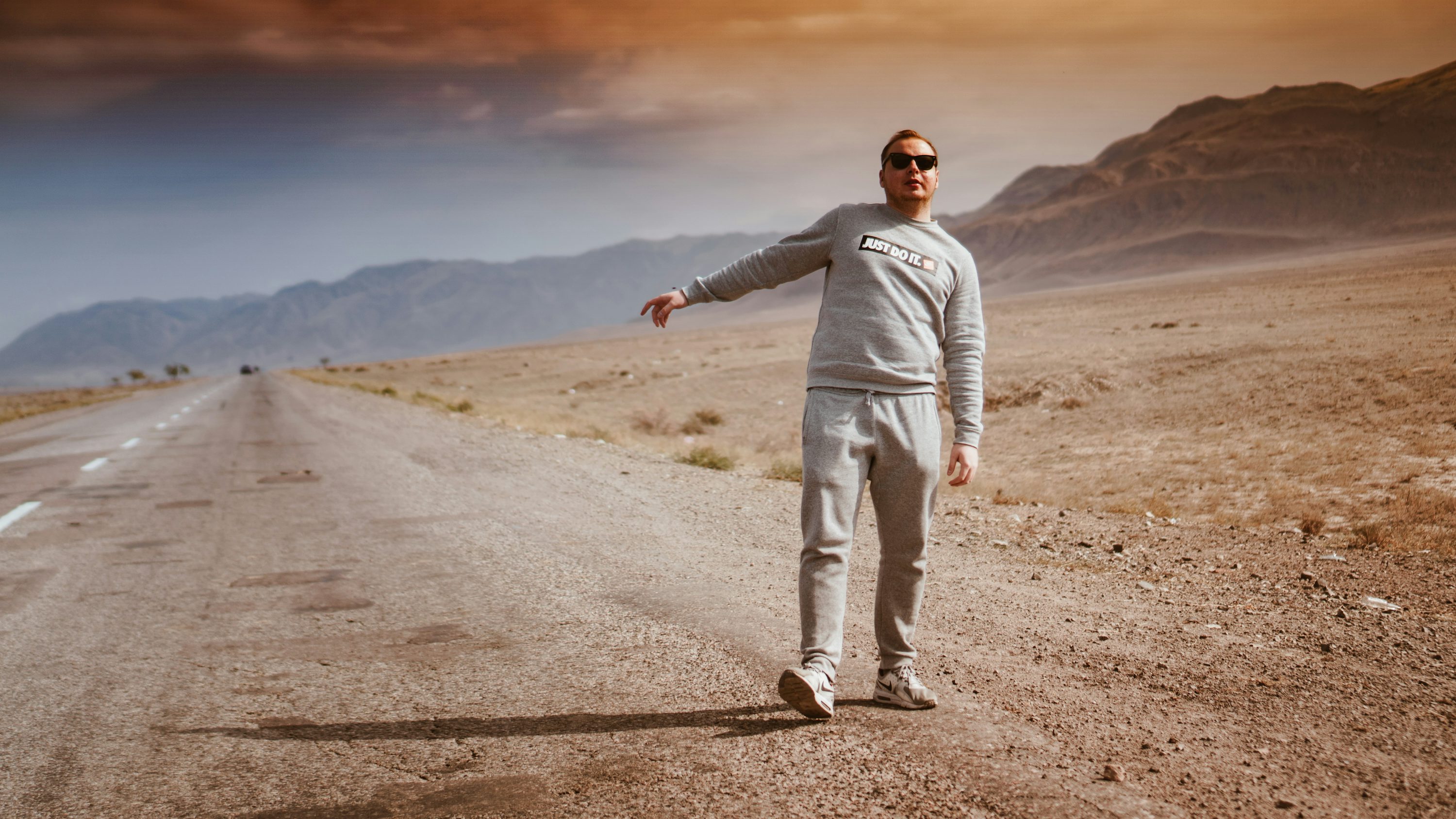 a man wearing a grey sweatshirt and sweatpants, sunglasses stands on a desert road, with his arm outstretched as if hitchhiking.  the road stretches into the distance, with mountains visible on the horizon. the scene is sunny and the landscape is arid.