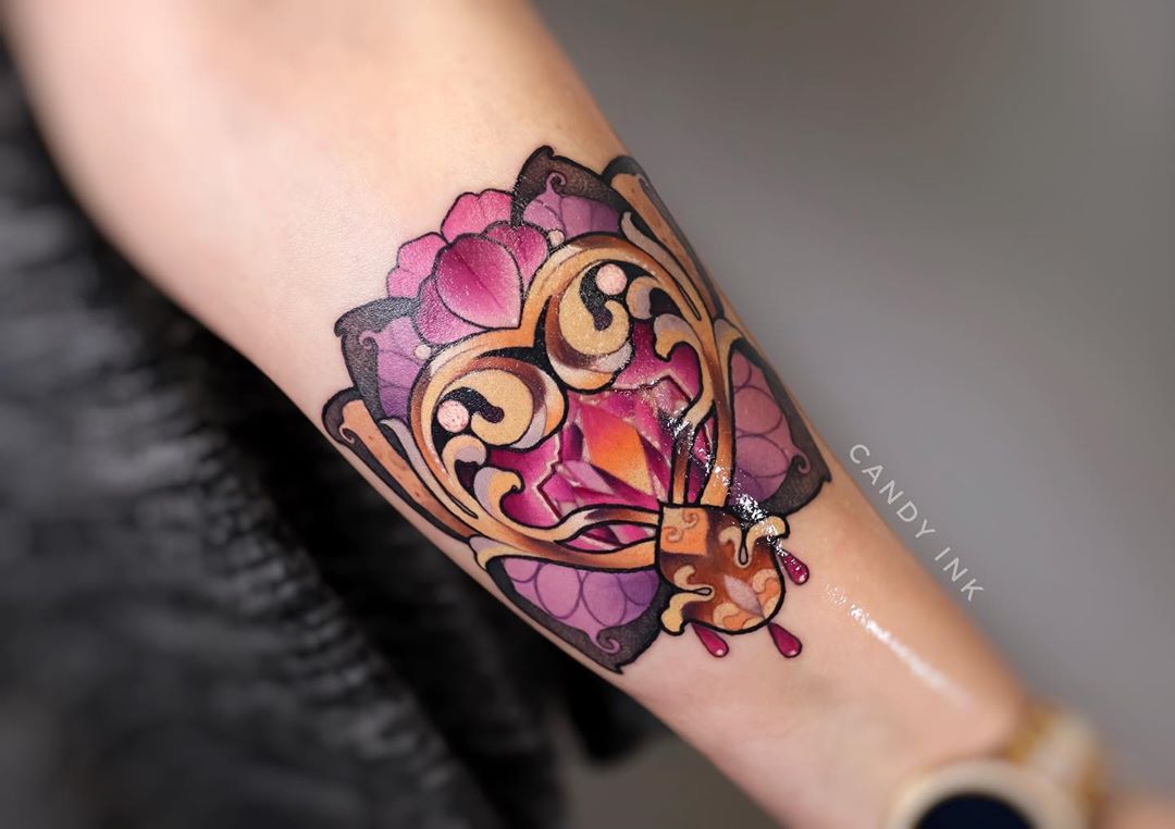 a closeup photo shows a neotraditional tattoo of an ornate golden heart with a pink and yellow jewel in the center, surrounded by pink and purple floral designs.  the tattoo is on someone’s inner forearm, just above their wrist.  there are three drops of red “blood” coming from the bottom of the heart. the photo is taken from above, looking down on the tattoo. the background is out of focus, but the tattoo is clear and sharp. the colors in the tattoo are bright and vibrant.
