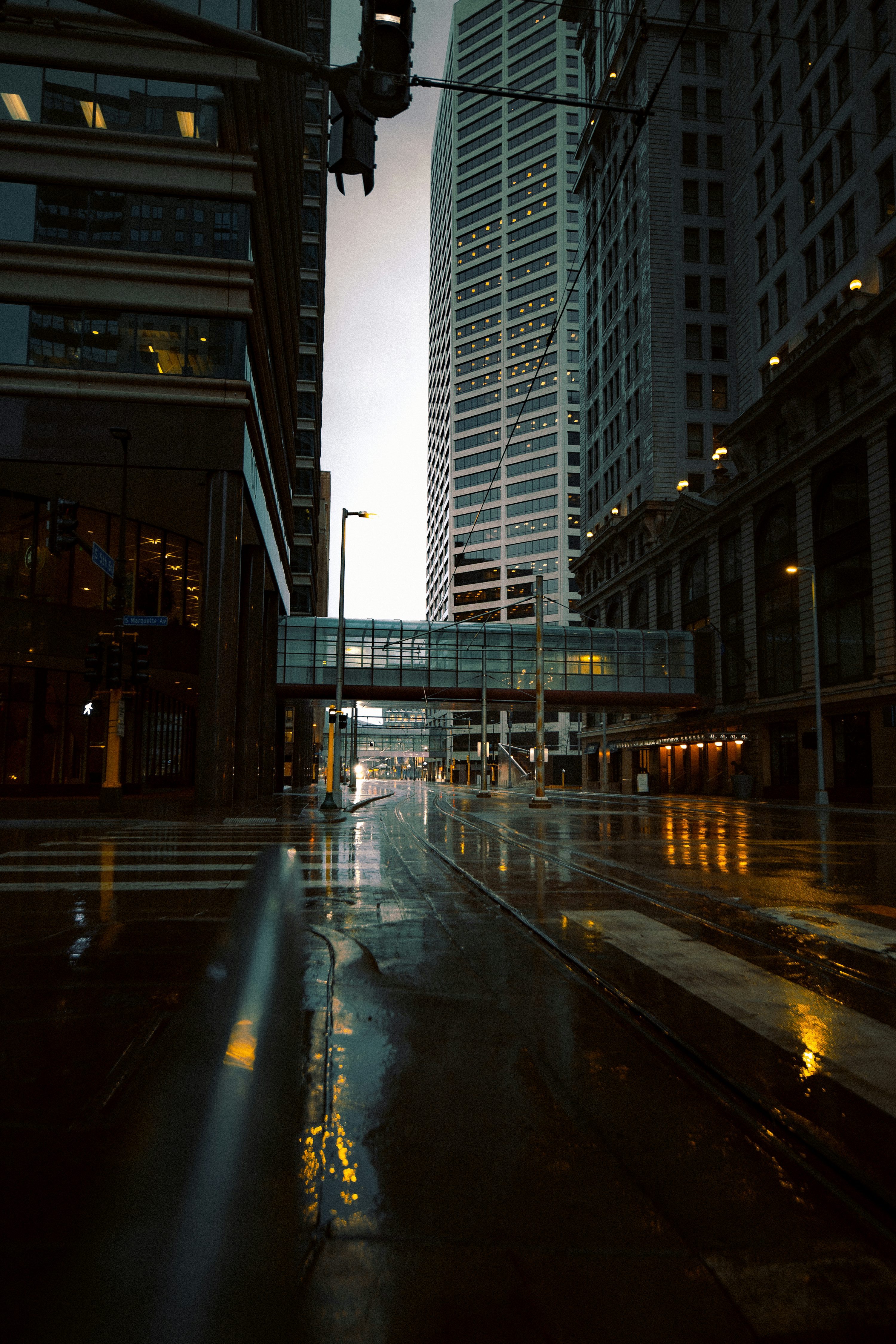 an urban street scene on a rainy evening. the wet pavement reflects the lights from the buildings and street lamps. skyscrapers dominate the background, connected by a skyway. a traffic light hangs above the street.