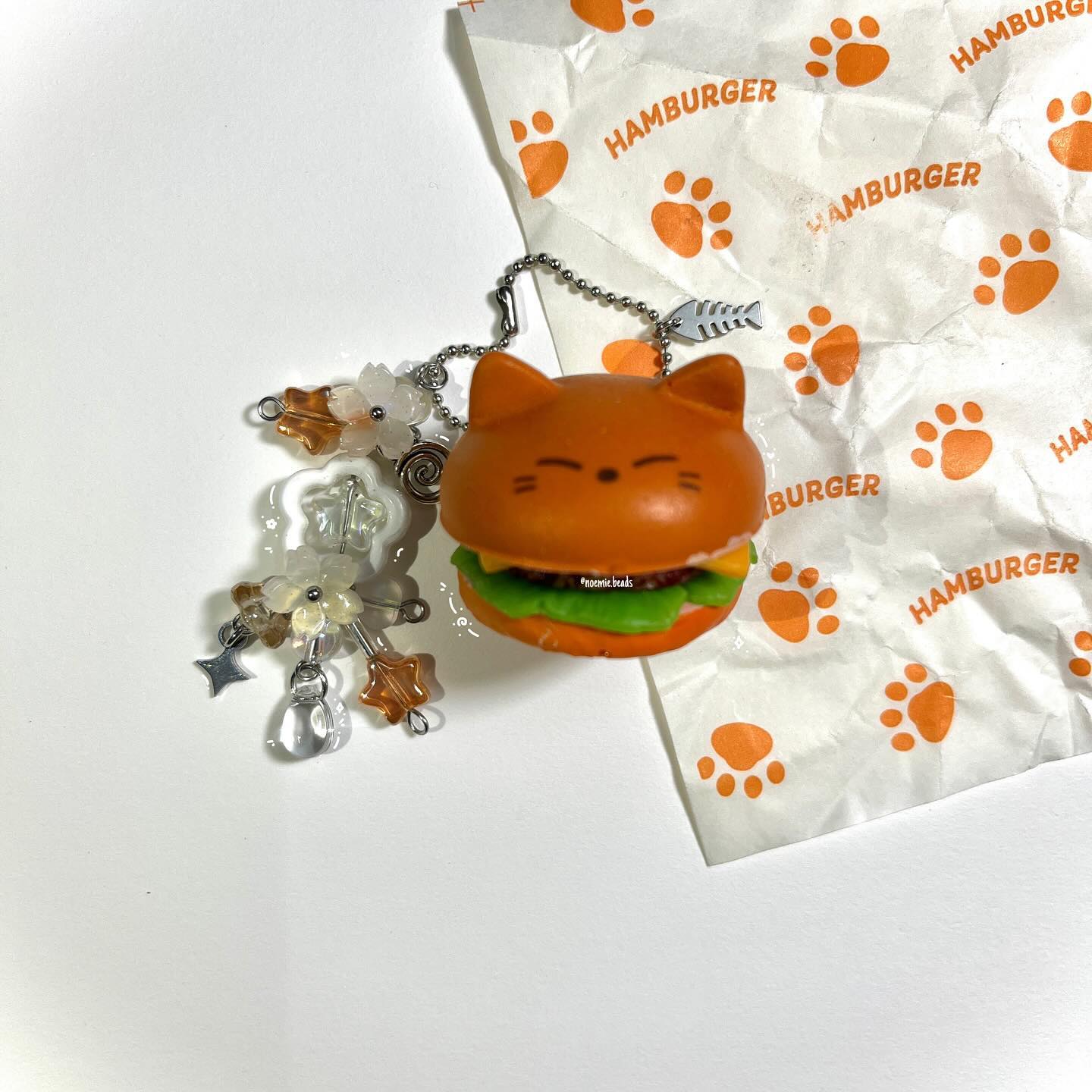 a cat burger keychain with charms is laying on a white surface next to a hamburger wrapper. the keychain features a cat head burger with green lettuce and a metal chain with a fish charm. there are also beaded charms with flowers and stars attached to the keychain. the hamburger wrapper has orange paw prints and the word "hamburger" printed on it.