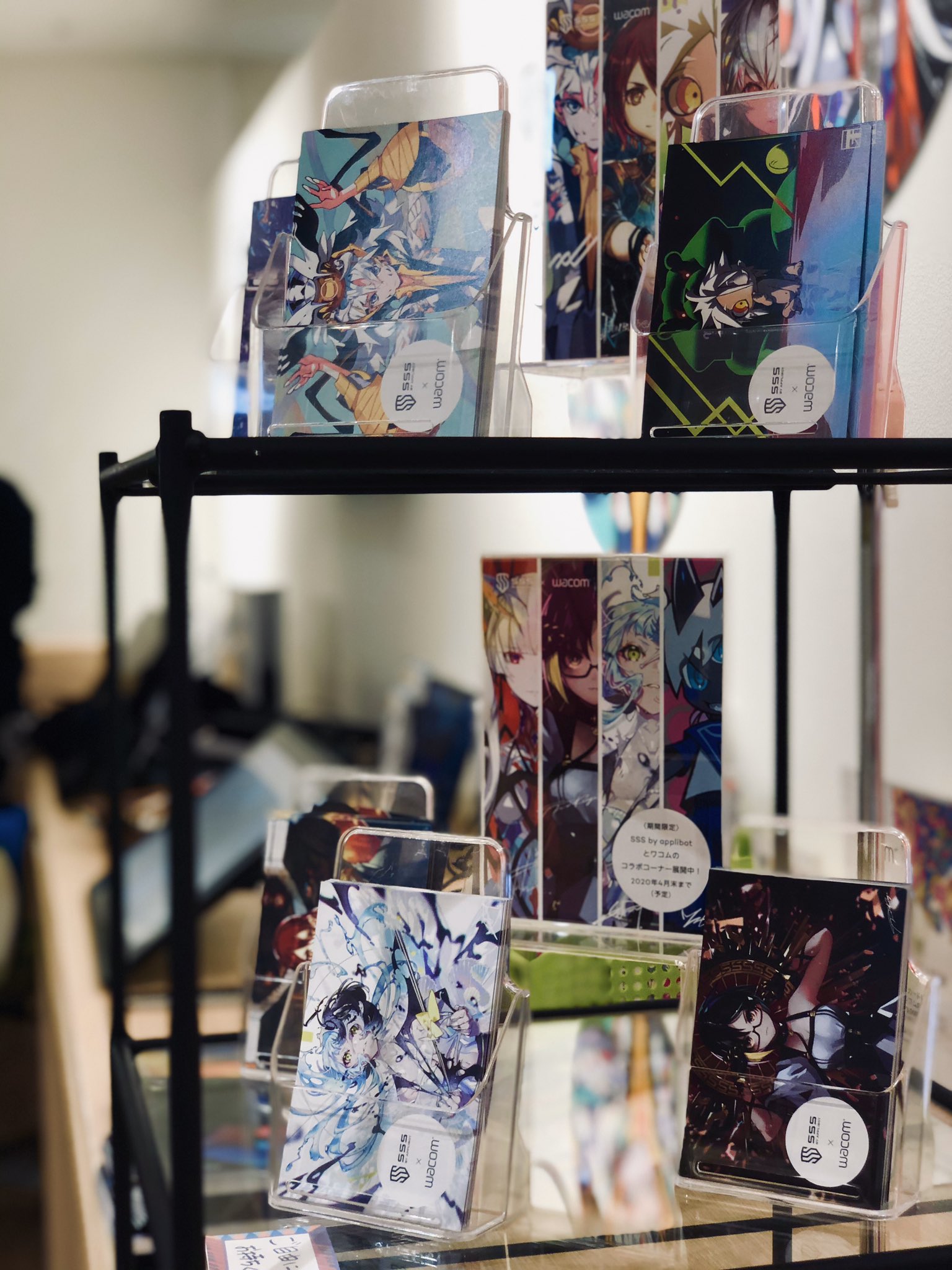 the image shows a black metal display rack with several anime art cards in it. the cards feature colorful illustrations of anime characters and are arranged in a way that showcases their designs. the rack is likely located in a store that specializes in anime merchandise.