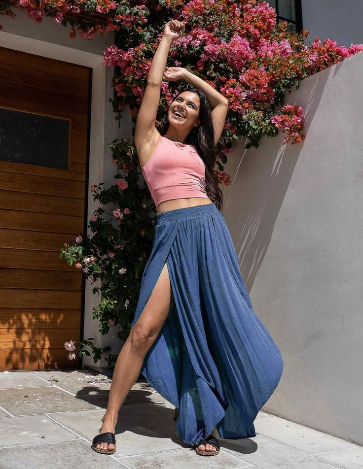 a young woman with long dark hair is posing outdoors in front of a flowering vine. she's smiling and wearing a pale pink crop top and highwaisted, wideleg blue pants with a thighhigh slit. her outfit is summery and stylish. she's wearing black slide sandals and is lit by natural sunlight, giving the image a bright and cheerful feel. the background features a brown door and stone tiles.