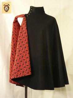 a black wool cloak with a red lining is shown on a mannequin. the cloak is open in the front to show the lining. the cloak is fastened at the neck with a single button. the lining is a red and black floral pattern. the cloak is floor length. the image is taken indoors in a studio setting. the lighting is artificial. the cloak is the focus of the image.