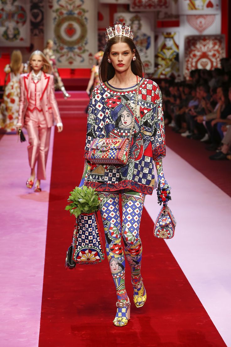 a model walks the runway during a dolce & gabbana fashion show.  she's wearing a vibrant, patterned outfit featuring playing card motifs, a crown, and colorful tights.  she carries several bags and her overall look is elaborate and theatrical, reflecting a playful, perhaps alice in wonderlandinspired theme. the runway is red and the background includes various playing card imagery.  another model in a pink suit can be seen further back.