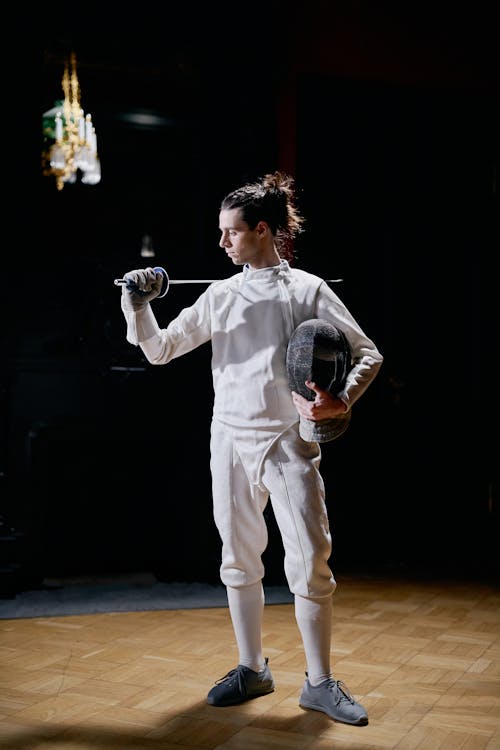 a young male fencer, dressed in a white fencing uniform, stands on a wooden floor in a dimly lit room. he holds a foil in one hand and a fencing mask under his arm.  his posture suggests concentration and readiness. the background is dark and slightly out of focus, drawing attention to the athlete. the overall mood is one of elegance and athleticism.