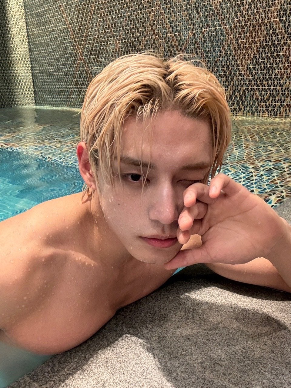a young  man with wet blonde hair is taking a selfie in an indoor swimming pool. he's shirtless and is resting his head on his left hand while looking directly at the camera. the pool has blue tiles.