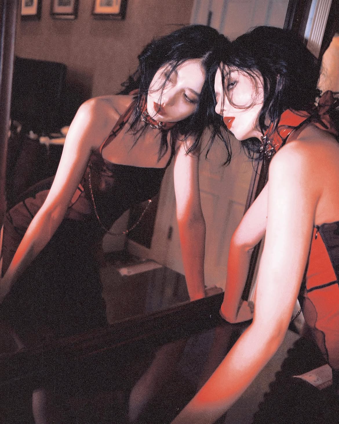 a woman with dark hair and red lipstick stands in front of a mirror. she is wearing a dark dress with a chain necklace and a choker. the mirror reflects her image.