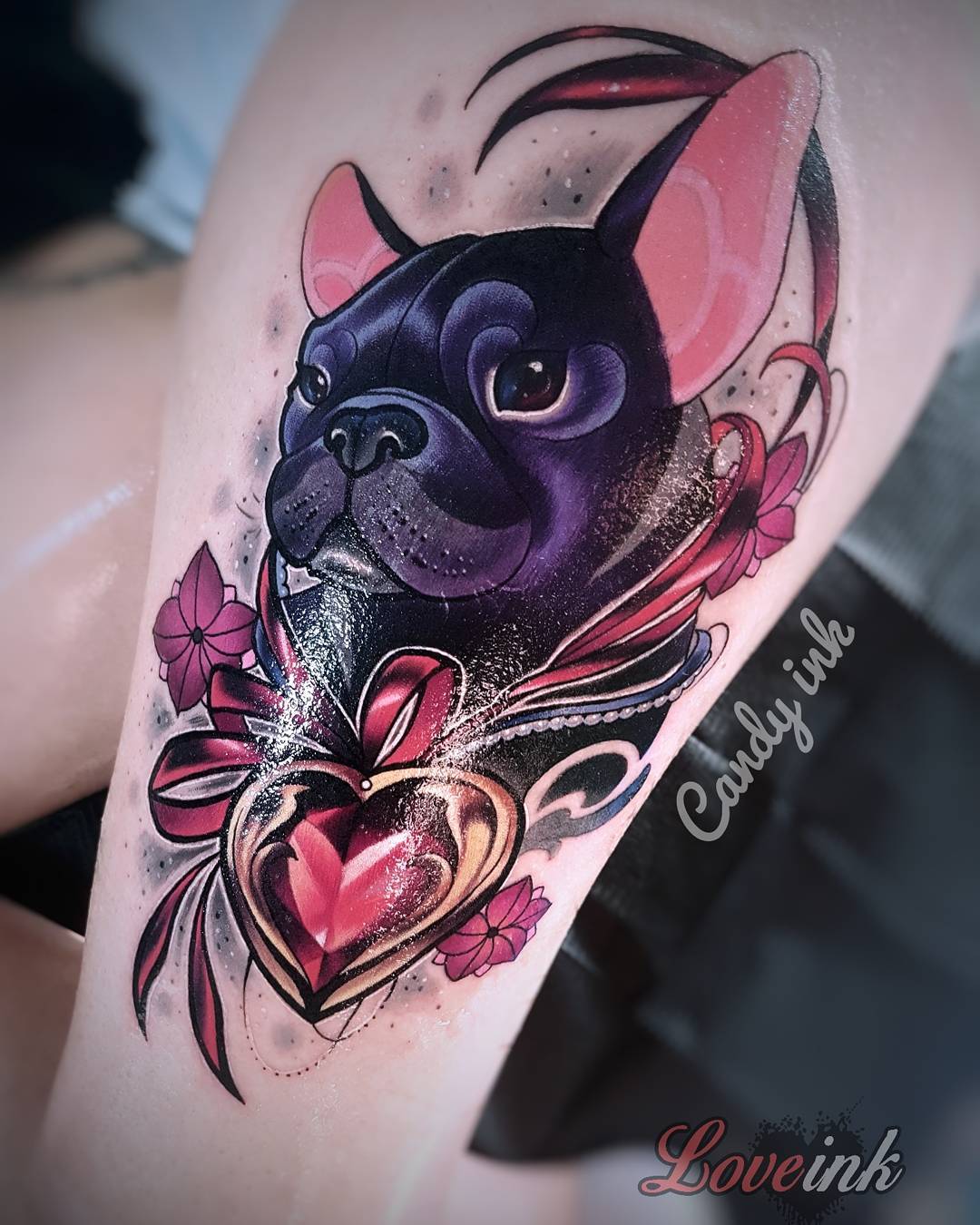 a neotraditional tattoo on someone's upper thigh. the tattoo features a black and grey french bulldog wearing a pearl necklace with a red and gold heart pendant.  there is a pink ribbon tied above the heart and pink flowers on either side of the dog's face.  the tattoo was done by candy ink and love ink.