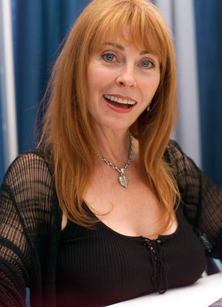 a woman with long red hair is smiling, her red hair is framing her face. she's wearing a black tank top with a lace overlay. she's also wearing a silver necklace with a heart pendant. the photo is taken in a casual indoor setting. the woman is looking directly at the camera and has a bright, friendly expression.