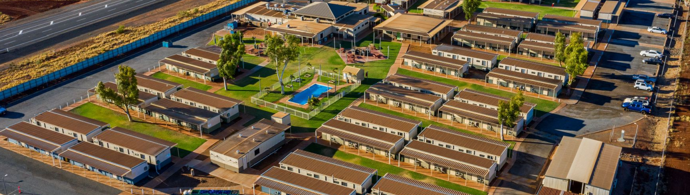 an aerial view of a motel complex in the outback of australia. the complex is made up of numerous cabins and bungalows with brown roofs and white walls, surrounded by green grass and landscaping. there is a swimming pool in the center of the complex. the parking lot is full of cars. the surrounding area is desert.