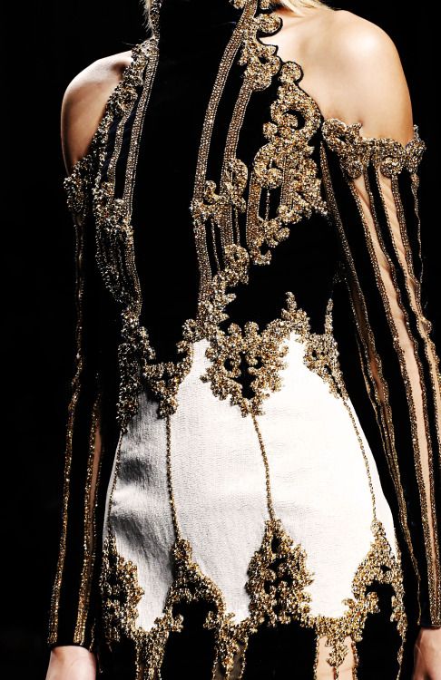 the image showcases a closeup of a woman's upper body wearing a stunning highfashion evening gown.  the dress is predominantly black with a white underlay showing through the intricate detailing. it features elaborate gold beading and embroidery forming ornate patterns across the black fabric. the neckline is a oneshoulder design, with the sleeve showcasing the same intricate gold embellishment as the bodice. the style is luxurious and evokes a sense of vintage glamour. the model's skin and the details of the dress are beautifully highlighted, giving an overall impression of opulence and refined style. the background is plain black, drawing all attention to the exquisite dress.