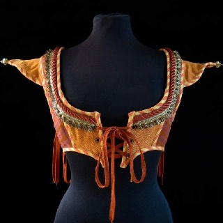 the image shows an orange and red bodice or corset on a black mannequin. the bodice features a laceup front with orange ribbon and is embellished with decorative trim, tassels, and ornamental details along the neckline and edges. the fabric has a textured appearance. the bodice has short, pointed sleeves. the background is plain black.