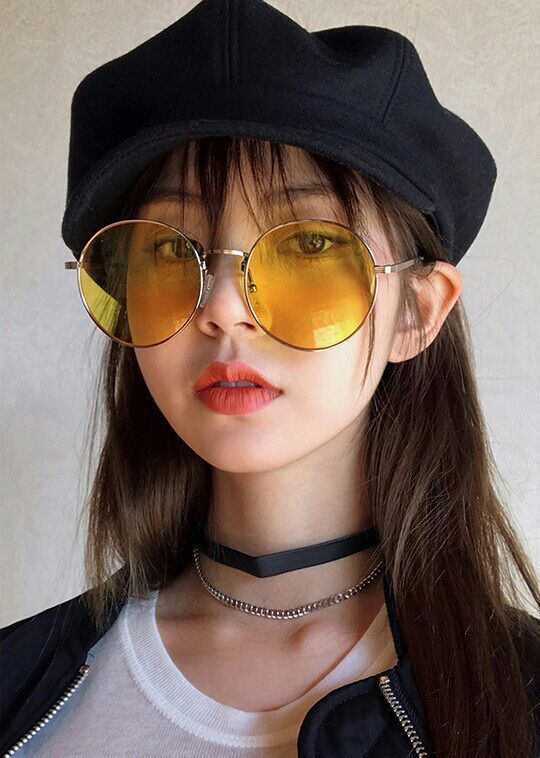 closeup portrait of a young woman with long brown hair wearing a black beret, yellow round sunglasses, and layered necklaces (a black choker and a silver chain). she is looking at the camera and has a stylish look.  the background is a muted gray.