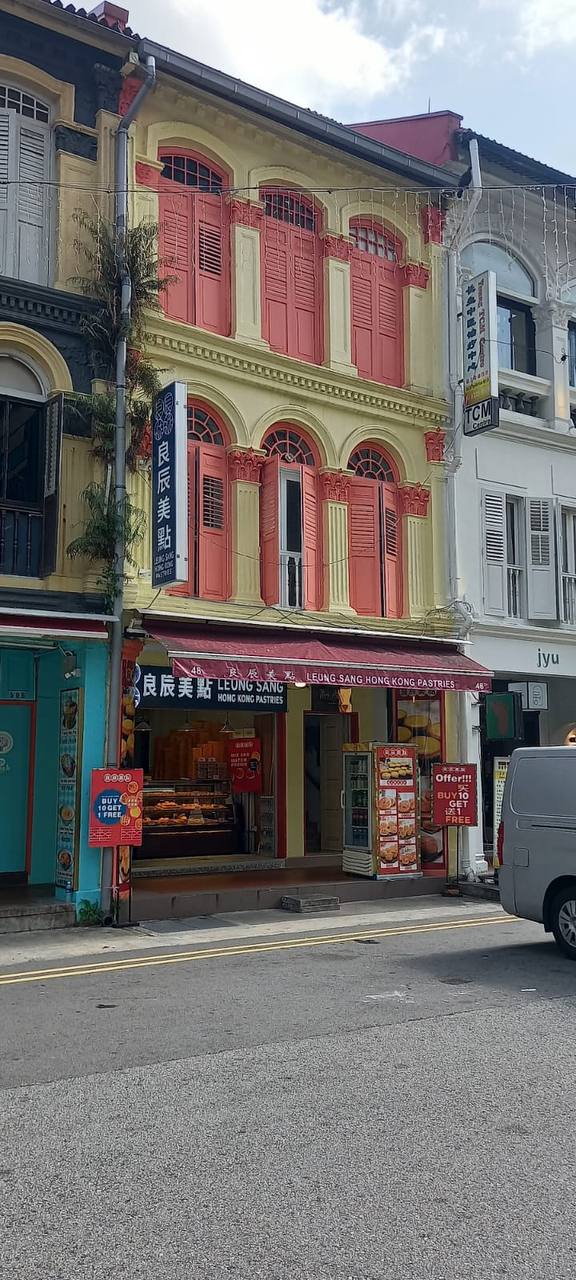 a streetlevel view of a traditional shop in hong kong. the building is brightly colored and decorated with ornate windows and shutters. the shop has an awning above the entrance and a sign that reads "leung sang hong kong pastries."  a white van is parked in the street.