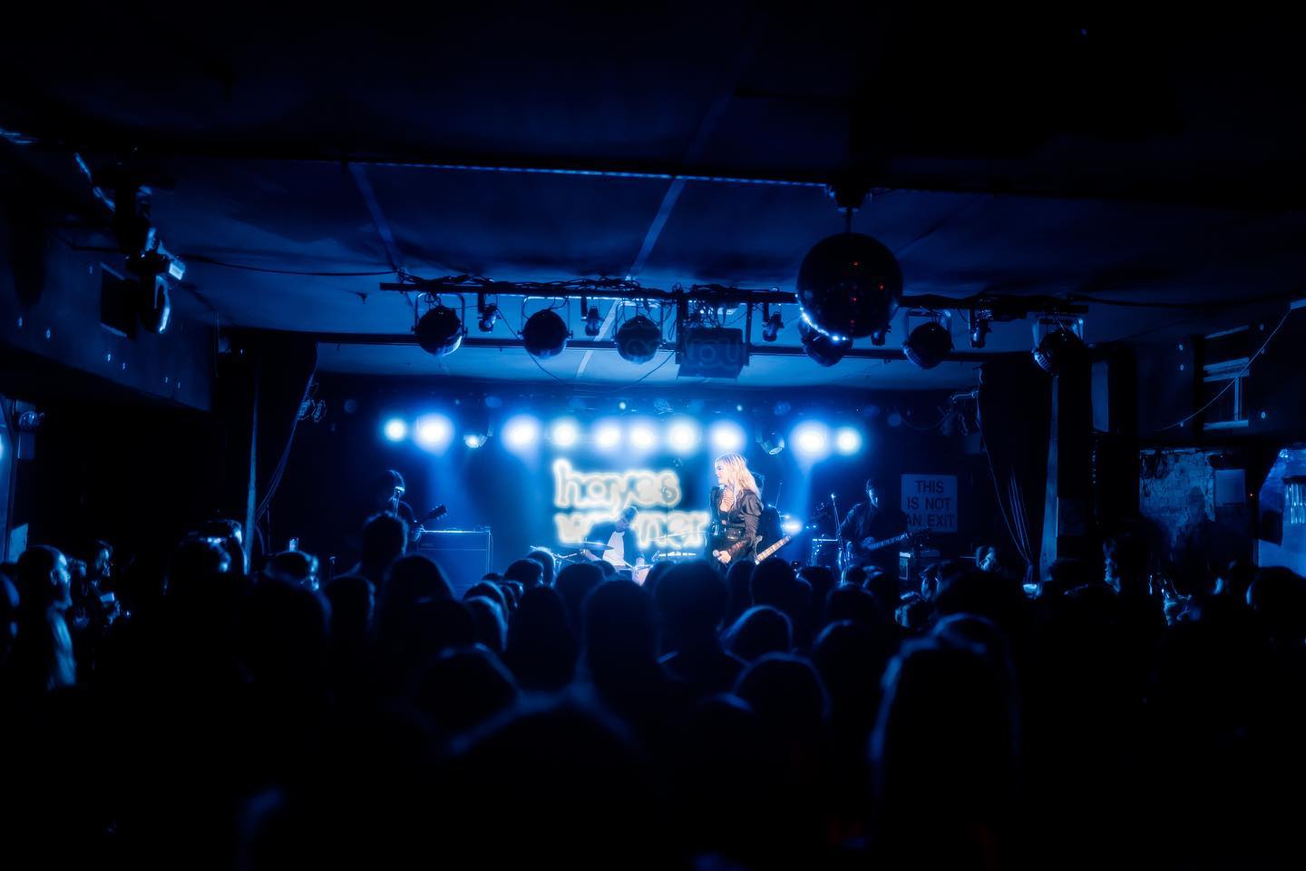 a concert taking place at a music venue, with the band "hayes warner" performing on stage under blue lighting. a crowd of people is in attendance, silhouetted against the light from the stage. disco balls hang from the ceiling. musicians are playing instruments including guitar and drums. a neon sign with the band's name is illuminated behind the singer.