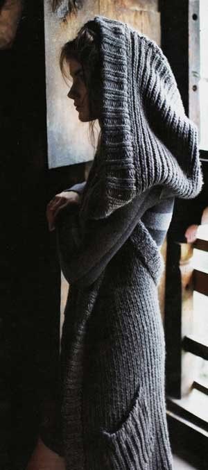 a woman wearing a long knitted gray cardigan, standing in front of a wooden door with a woodpaneled window. the woman has the hood of the cardigan up over her head. her head is slightly turned, looking over her shoulder, looking towards the left side of the image.  her eyes are not visible. the image is taken from a low angle, below the woman's head.  the background is out of focus.