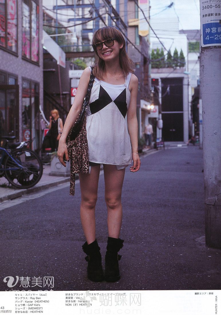 a young woman with shoulderlength brown hair and sunglasses is standing on a city street, posing for a fashion photo. she is wearing a white tank top with a black and white design, denim shorts, black ankle boots, and carrying a leopardprint handbag. she is smiling and looking at the camera. there is a bicycle parked on the sidewalk behind her, and buildings and signs are visible in the background.