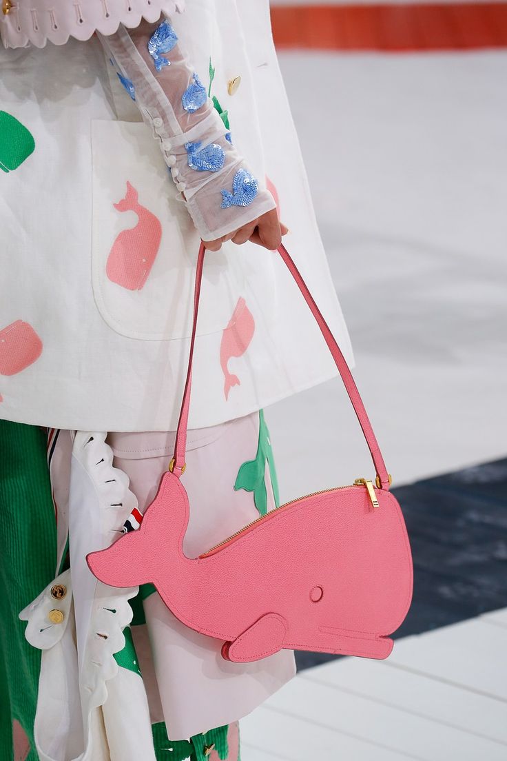 loseup of a model's outfit on a runway, showcasing a pink whaleshaped handbag. the bag is a light pink color and has a gold zipper. the model is wearing a white outfit with pink whale appliqués and blue sequined sleeves. the background is blurred, focusing on the bag and outfit details. the overall style is whimsical and playful, suggesting a spring or summer collection.
