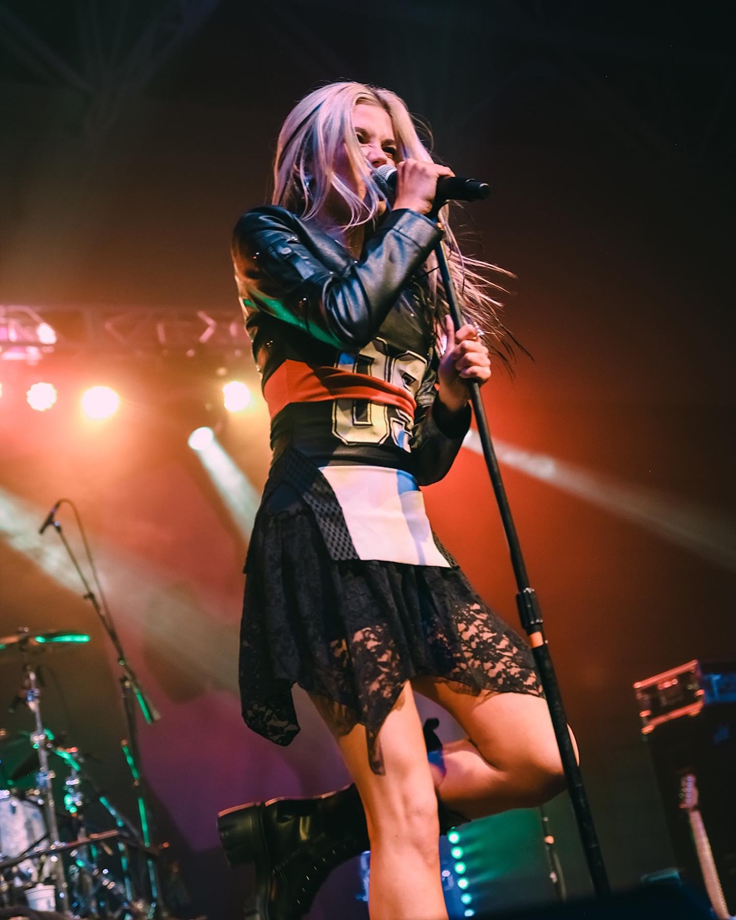 a woman is singing into a microphone on a stage during a concert. she has blonde hair and is wearing a leather jacket, a dress, and boots. there are drums and a guitar visible in the background. the stage is lit with colorful lights.