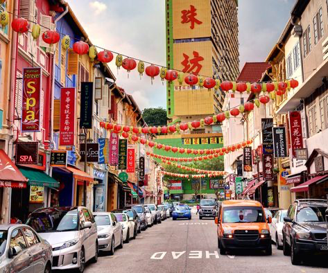 the image is a vibrant street scene in singapore's chinatown, likely during chinese new year.  rows of brightly colored shop houses line a narrow street, adorned with numerous red and yellow lanterns strung overhead. the architecture is traditional, with various businesses visible, including spas and massage parlors.  the street is filled with parked cars, adding to the bustling atmosphere. a variety of chinese and english signage adds to the cultural richness of the scene. the overall impression is one of lively energy and cultural vibrancy.