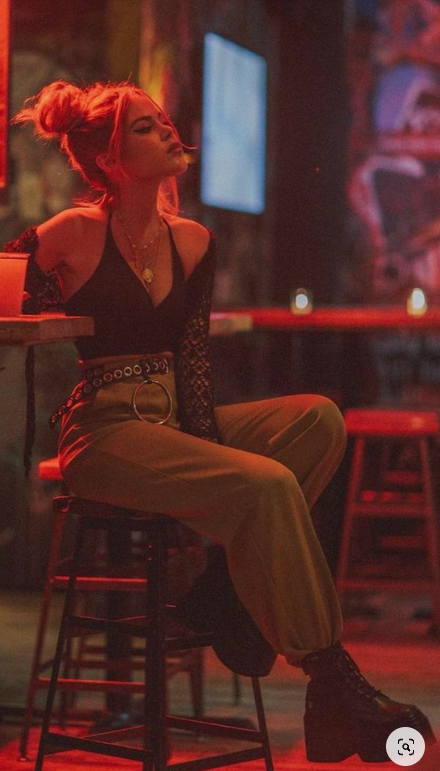 a woman with blonde hair in a bun is sitting on a bar stool in a dimly lit bar. she is wearing a black crop top, brown pants, and black platform boots. the lighting is red and casts a warm glow on the scene.