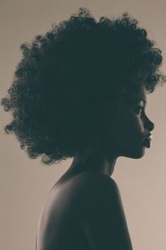 the image shows a woman of color with an afro hairstyle. she has dark skin and is facing to the right of the frame.  the background is a light brown color, and the image appears to be professionally lit.