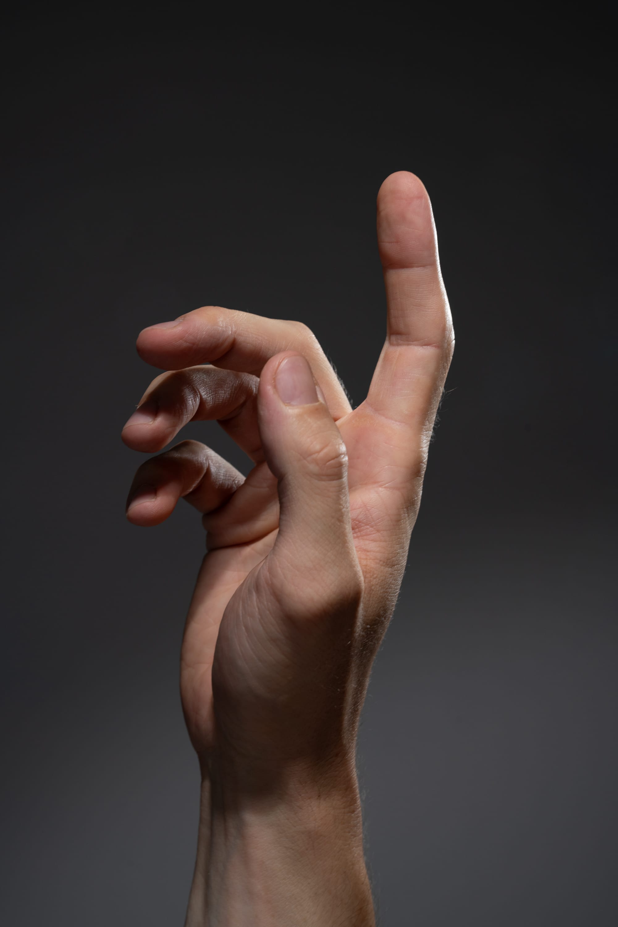 a man's right hand is extended against a gray background. the hand is positioned with the index finger pointing upwards and the other fingers slightly curled. the lighting is soft and directional, highlighting the texture and details of the hand.