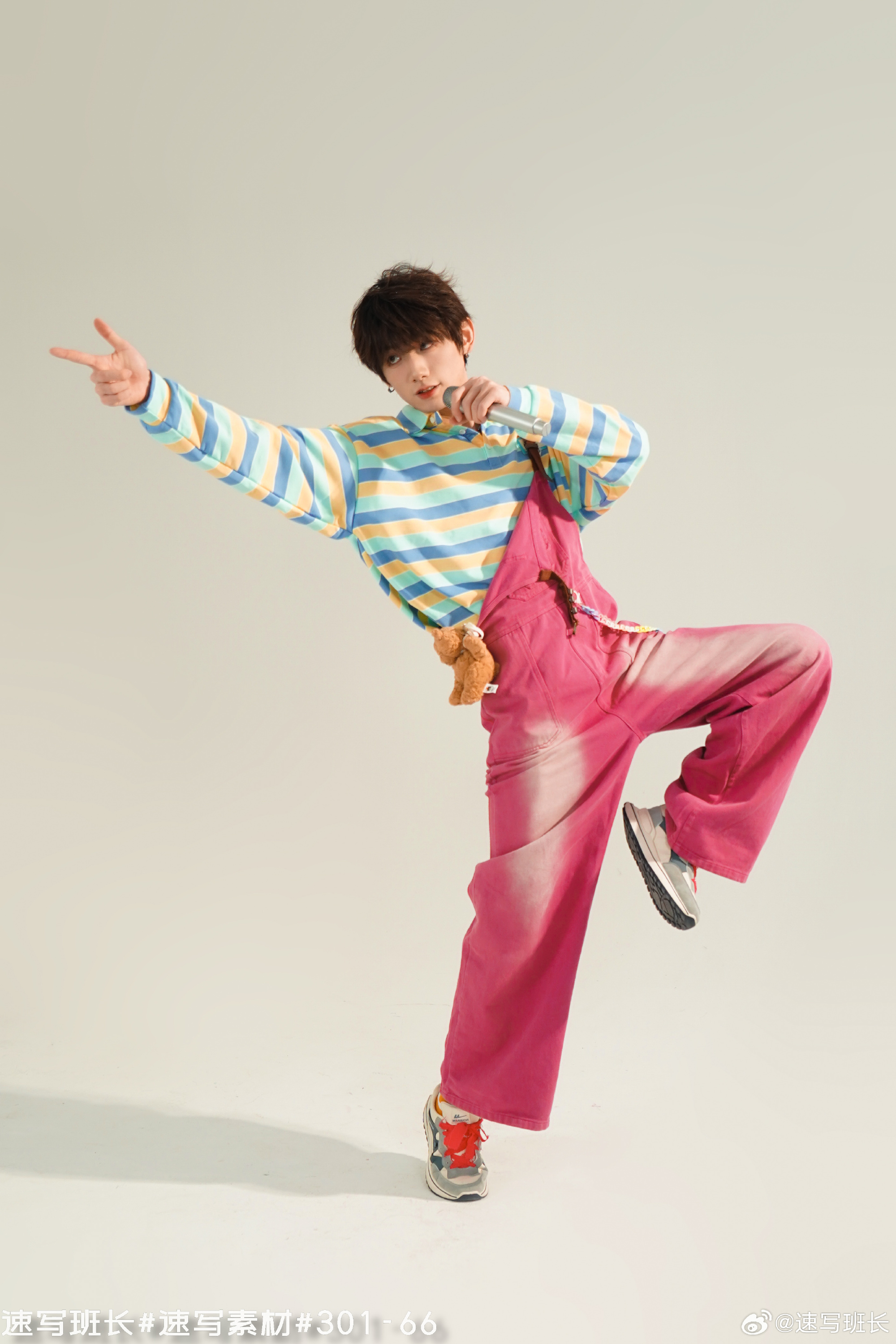 a young man in a colorful outfit is singing and dancing in a studio. he is wearing a striped sweater, pink overalls, and sneakers. he is holding a microphone in his right hand and making a peace sign with his left hand. there is a small teddy bear attached to his overalls. the background is white and the lighting is bright.