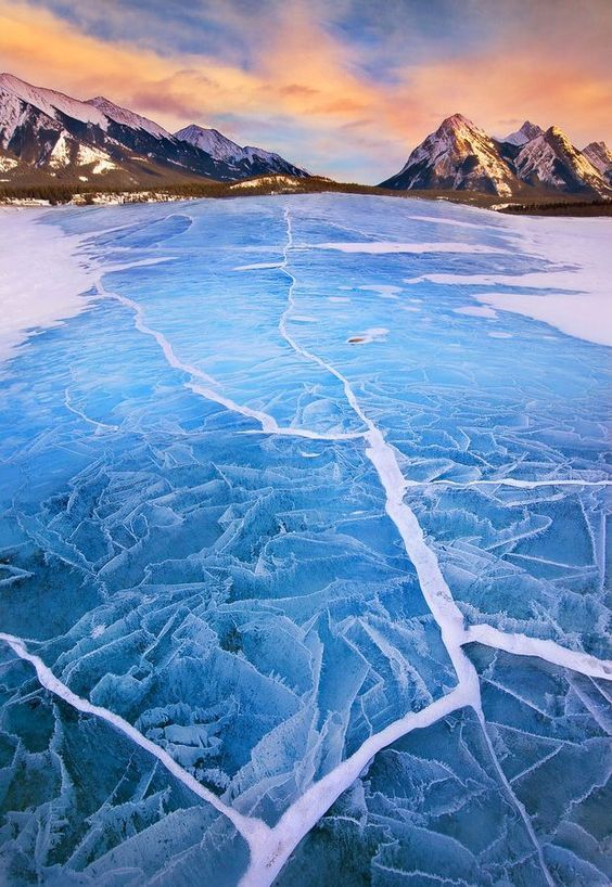 the image captures a stunning winter landscape featuring a frozen lake with a network of intricate cracks sprawling across its icy surface. in the background, a majestic mountain range is silhouetted against a vibrant sky painted with the warm hues of a sunset. the scene evokes a sense of serene beauty and the raw power of nature.