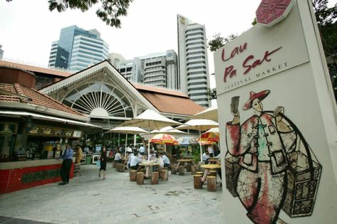 the image shows lau pa sat, a historic market and hawker centre in singapore.  the foreground features several food stalls under umbrellas, with people sitting at tables enjoying food.  the background shows the modern skyscrapers of the city. a large sign advertising "lau pa sat festival market" is prominently displayed.  the style of the architecture is a blend of victorian and asian influences. the image conveys a bustling, vibrant atmosphere of a city market.