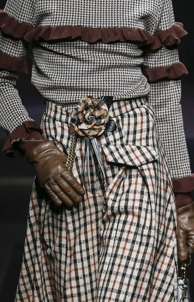 loseup view of a woman's fall/winter outfit. the outfit consists of a houndstooth patterned top with brown ruffled detailing at the cuffs and neckline.  a plaid skirt in cream, brown, and black is worn over the top. a large, brown floral brooch is fastened to the skirt at the waist, where a brown leather belt is also visible. the woman is also wearing brown leather gloves.  the image is a detail shot, focusing on the texture and design elements of the clothing. the setting appears to be a fashion show or photoshoot.