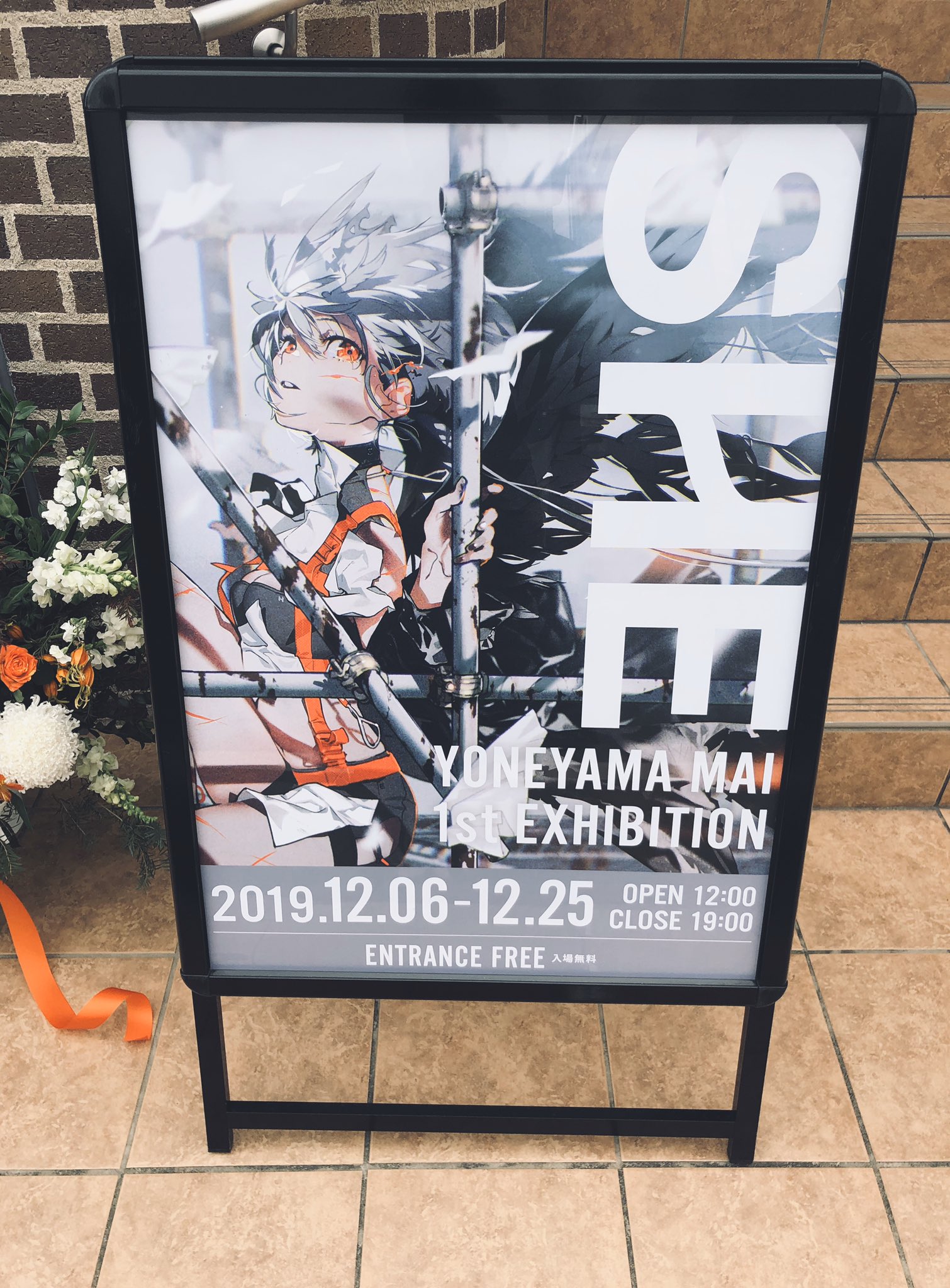 an aframe sign stands on a tiled sidewalk in front of a brick building, advertising the yoneyama mai 1st exhibition.  the sign features an animestyle illustration of a character with white hair and orange eyes partially obscured by black and orange scaffoldinglike lines.  the exhibition dates are listed as december 6th, 2019, to december 25th, 2019, with open and close times.  a bouquet of flowers is partially visible behind the sign.
