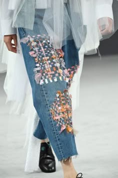 a closeup shot of a fashion model wearing a pair of denim jeans that are heavily embroidered with a patchwork of different textures and colors. the jeans are cuffed at the ankles and the model is wearing black boots. the image is taken from the waist down and the model is walking towards the camera.